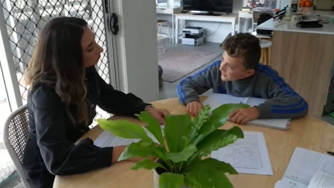 Clarissa sits at a table with Miles. Papers and books are spread out on the table.