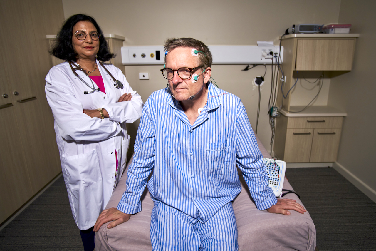 A woman wearing a white doctor's coat stands next to a man sitting on a bed in striped pyjamas 