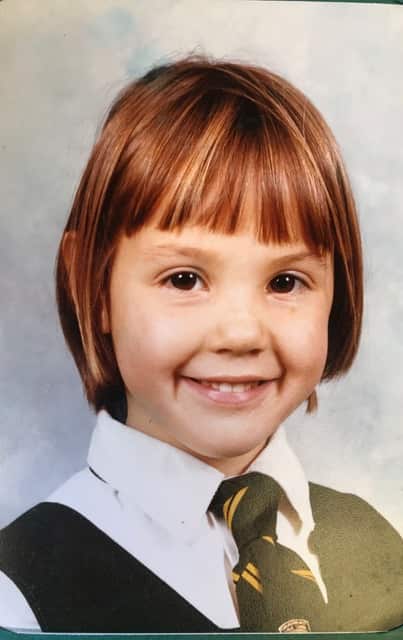 A portrait of a primary school girl with bobbed hair, who is smiling at the camera. She is wearing a school uniform tunic, white shirt and tie