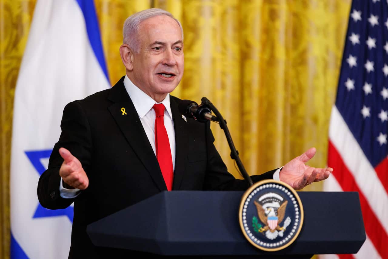 A man in a dark suit with a bright red tie stands behind a podium in front of the Israeli and US flags