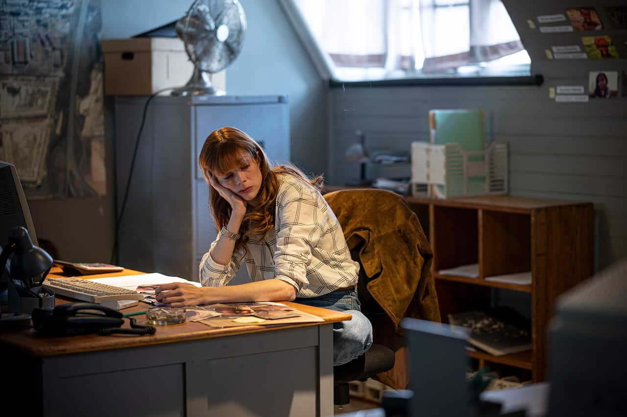 A woman sits at a desk in a shadowy office, with her hand resting on one hand.