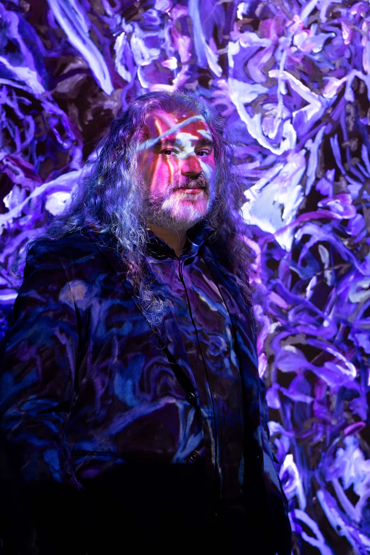 Michael Dagostino, a middle-aged Italian Australian man with long grey hair, stands in front of a vivid blue and purple painting being projected onto a wall.