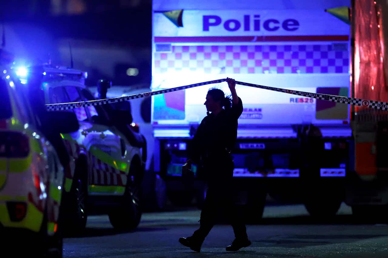 A police officer lifts crime-scene tape while walking between emergency vehicles illuminated by blue lights at night.