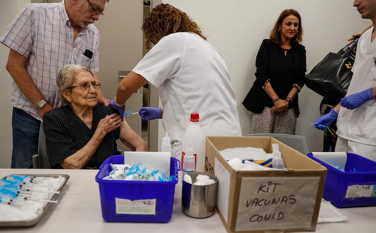 An older woman sitting in a chair. A nurse facing her to her right is giving her an injection in her left arm. There is a man standing behind her