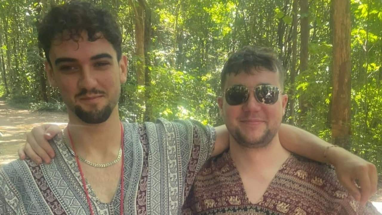 a closeup of two young men in patterned shirts with their arms around each other standing in a jungle