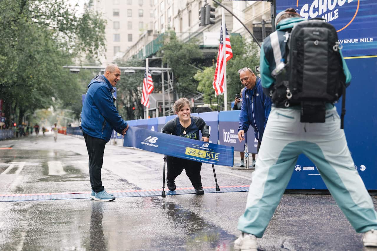 A woman of short stature is about to run through a blue ribbon with text reading '5th Ave Mile' on it.  She is using hand held crutches. Two men are holding the ribbon, and there is a third man pictured from the back.  