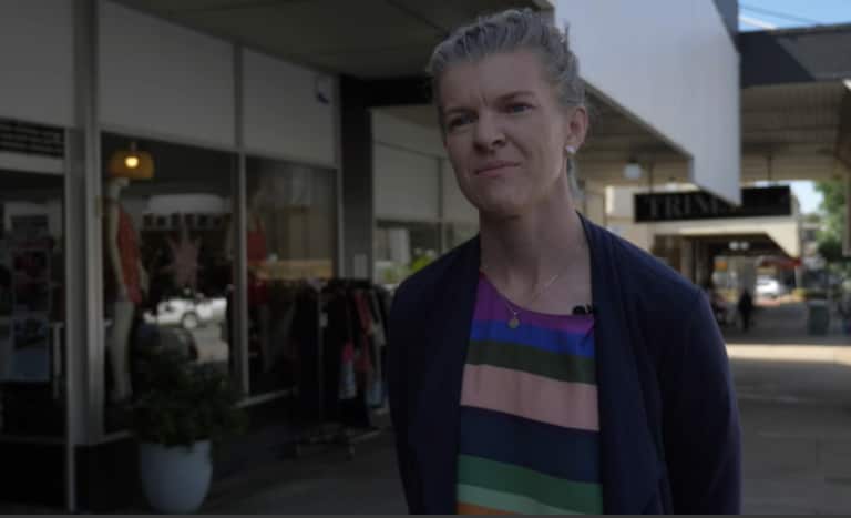 A middle-aged white woman with blonde hair, stands outside in front of several shopfronts.