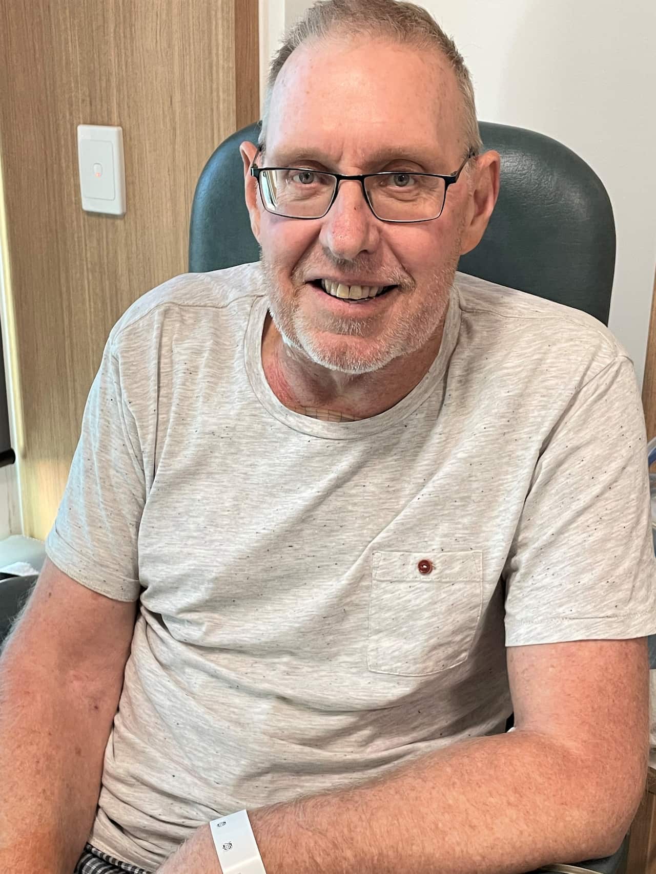 A man in a pale grey t-shirt sits smiling at camera. 