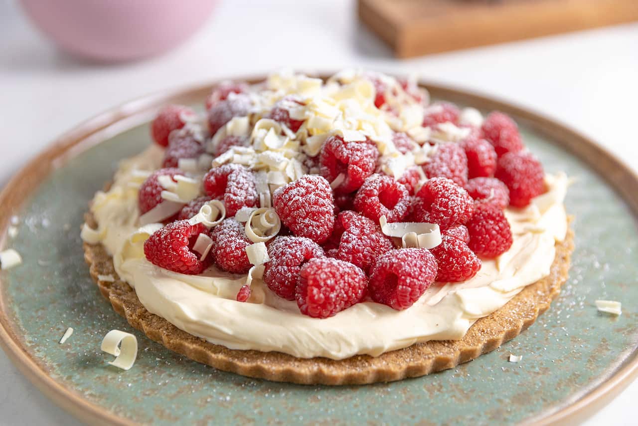 On a lovely wide, flat green-toned plate sits a disk of pastry, topped with a layer of whipped cream, fresh raspberries and white chocolate shavings.  