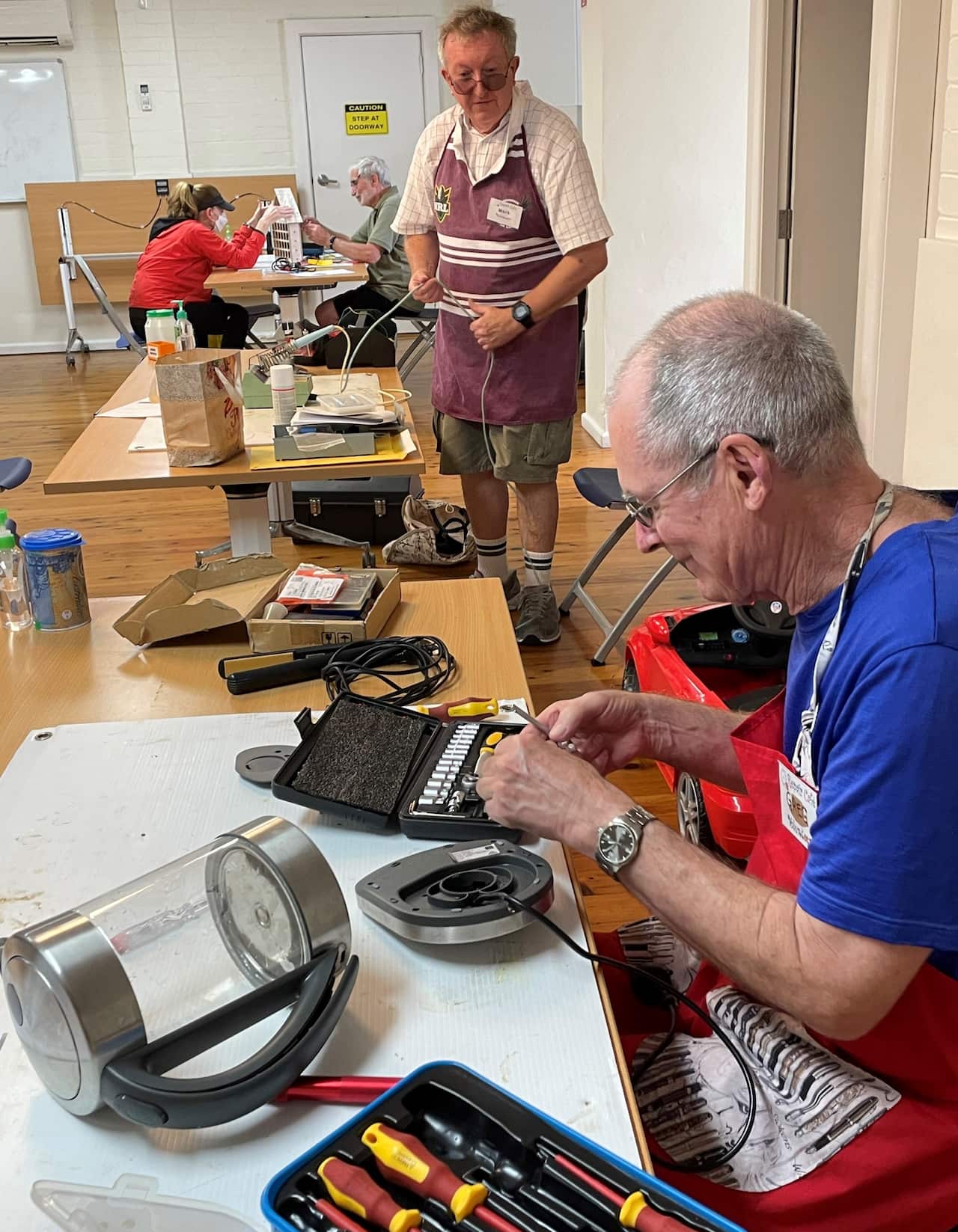 A man standing watches another man sitting at a table with electrical equipment.