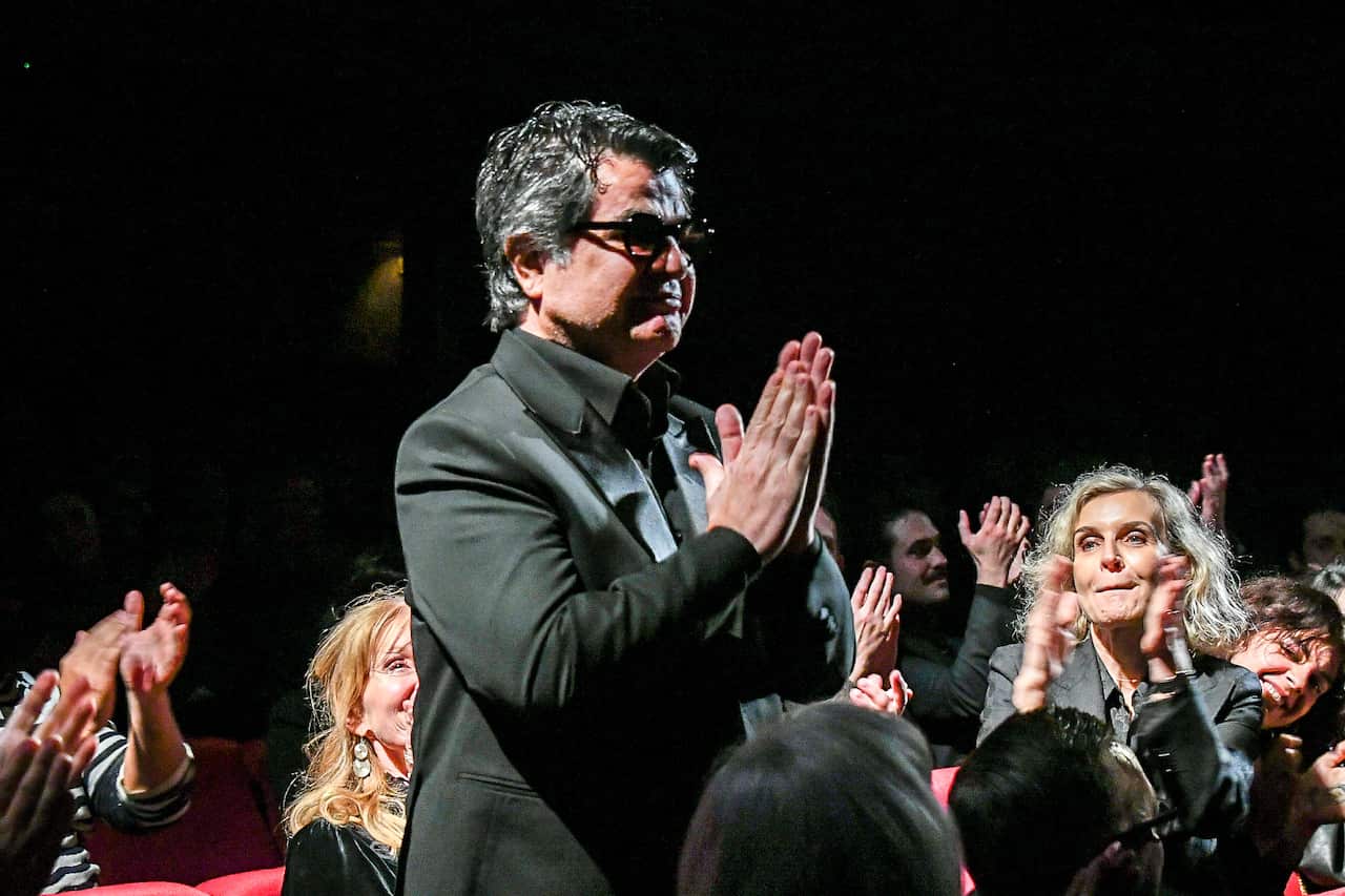 A man in an all-black outfit and sunglasses folds his hands as people clap around him.