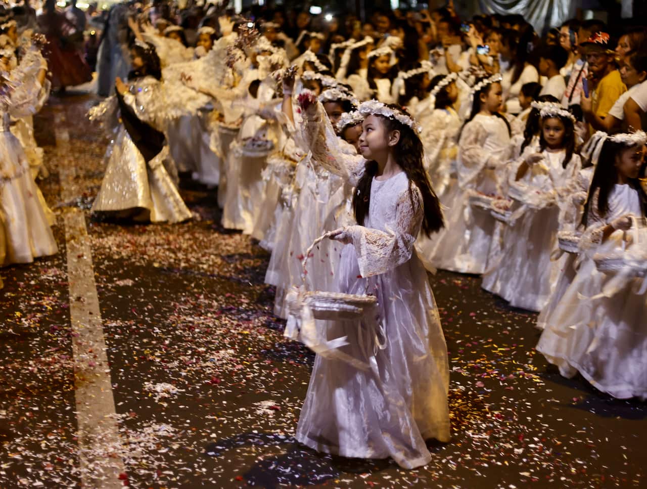 Young girls in white angel costumes scatter confetti while walking down a street together. 