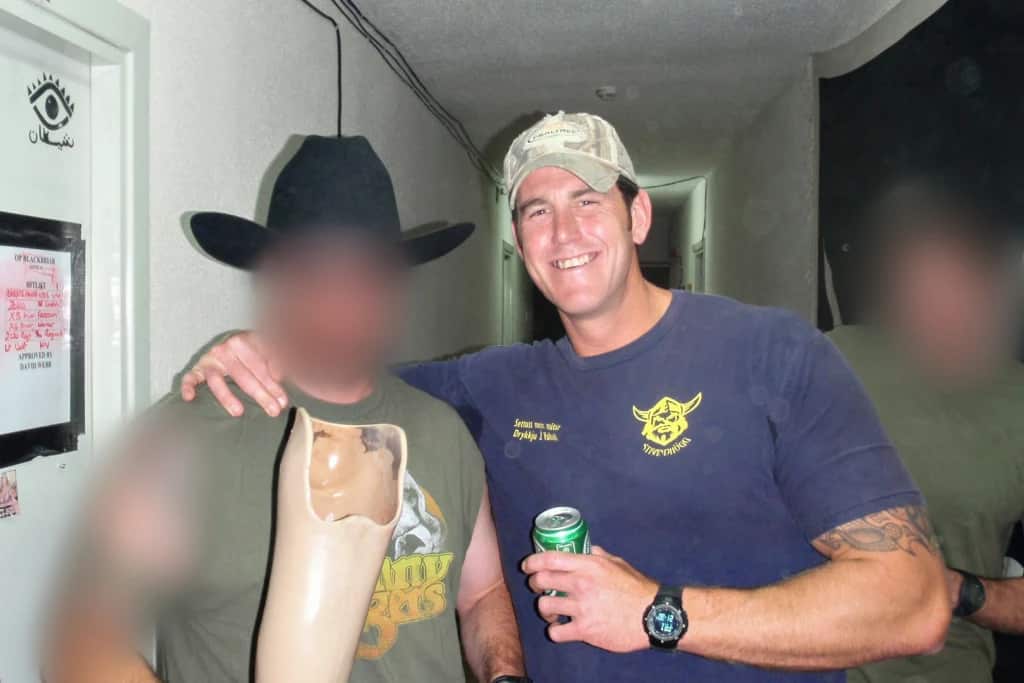 Ben Roberts-Smith drinking a beer as another man, blurred, holds a prosthetic leg.
