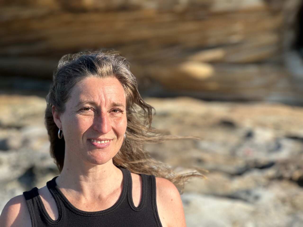 A woman with a black tank top and long brown hair smiles at the camera. It's a windy day and there are rocks in the background.