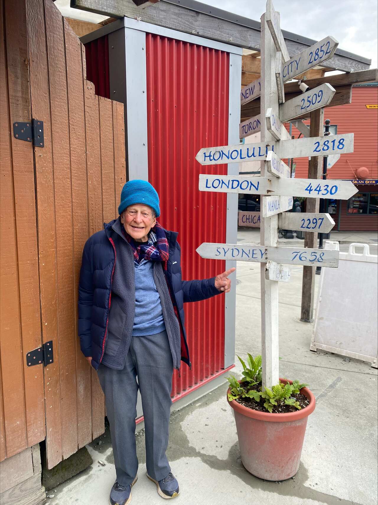 An elderly man standing next to a signpost that shows the distance to various cities around the world.  