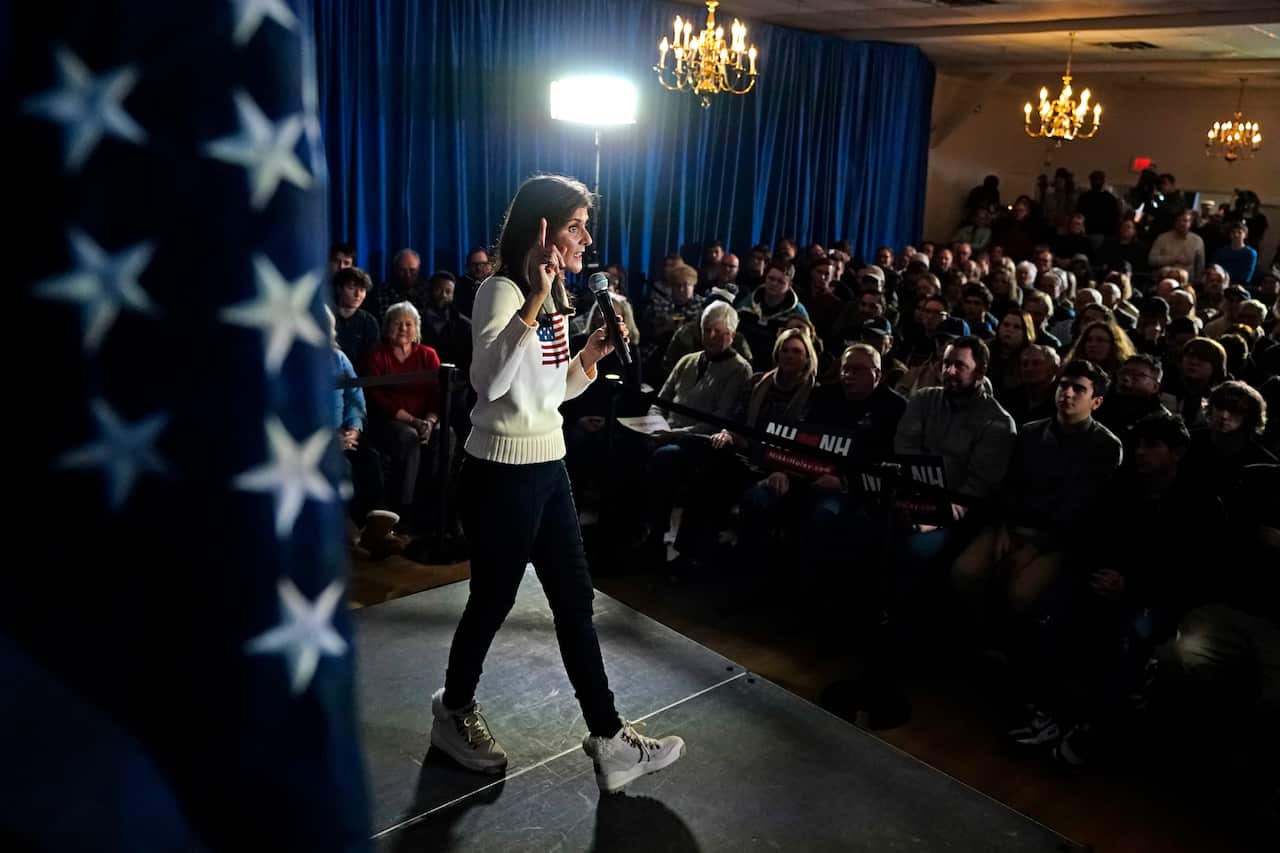 Woman in black jeans and white sweater with American flag speaks on stage, holding a microphone in front of a seated crowd.