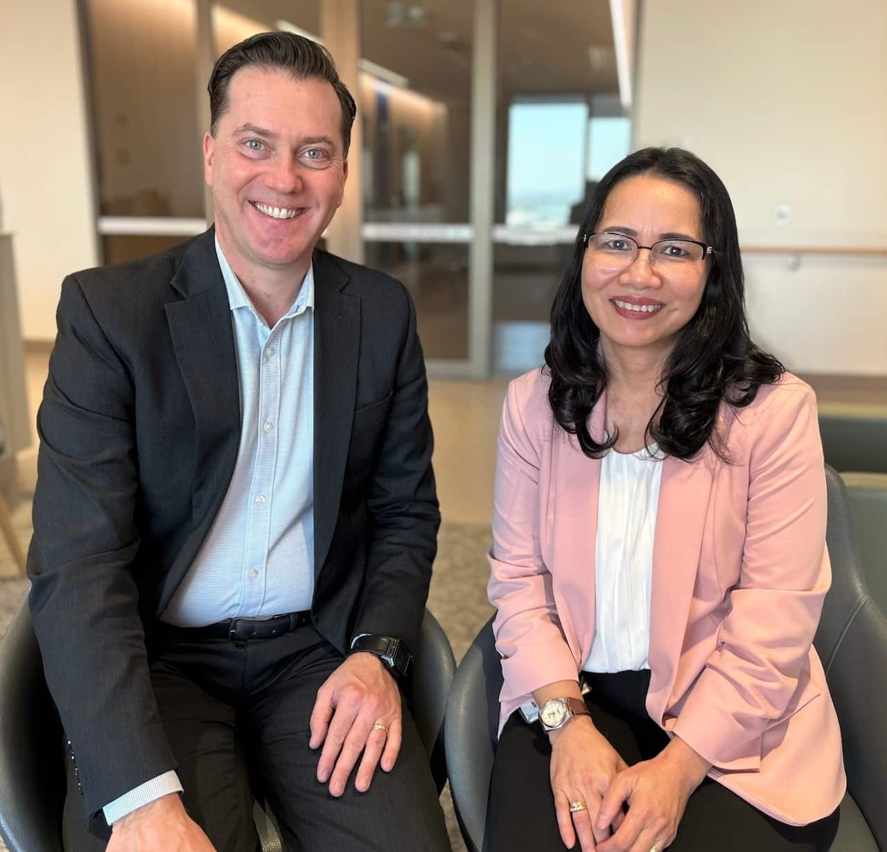 A man in a suit jacket sits with a woman in a pink jacket smiling at camera. 