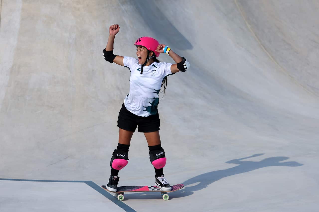 Arisa Trew riding a skateboard, wearing a pink helmet and kneepads, in a skatepark.