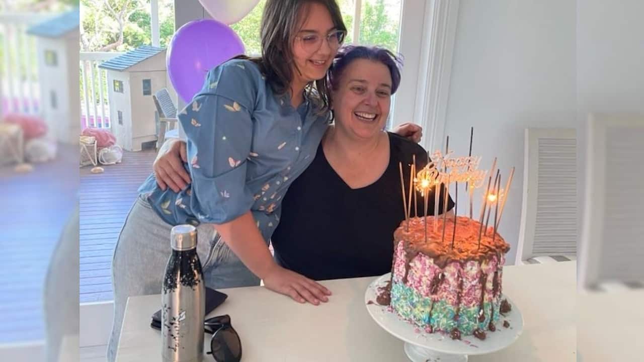 a young girl hugs a middle aged woman sitting at a kitchen bench with a birthday cake in front of them