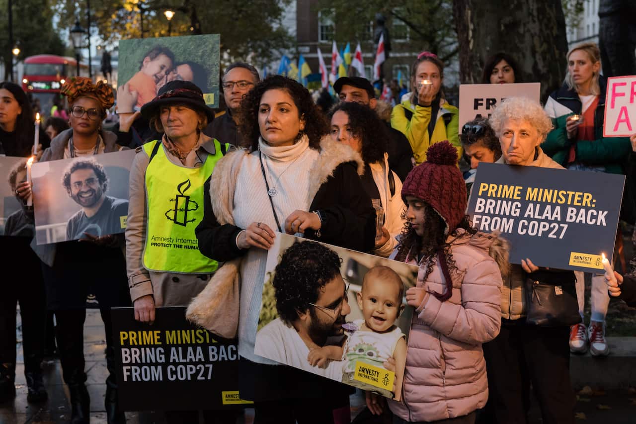 A group of people gathered at dusk holding candles and placards featuring the face of Alaa Abd el-Fattah.