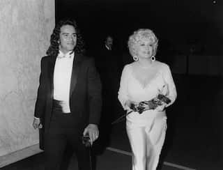 Black-and-white photo of two people in formal attire walking indoors, with one carrying a bouquet of roses and large earrings visible.