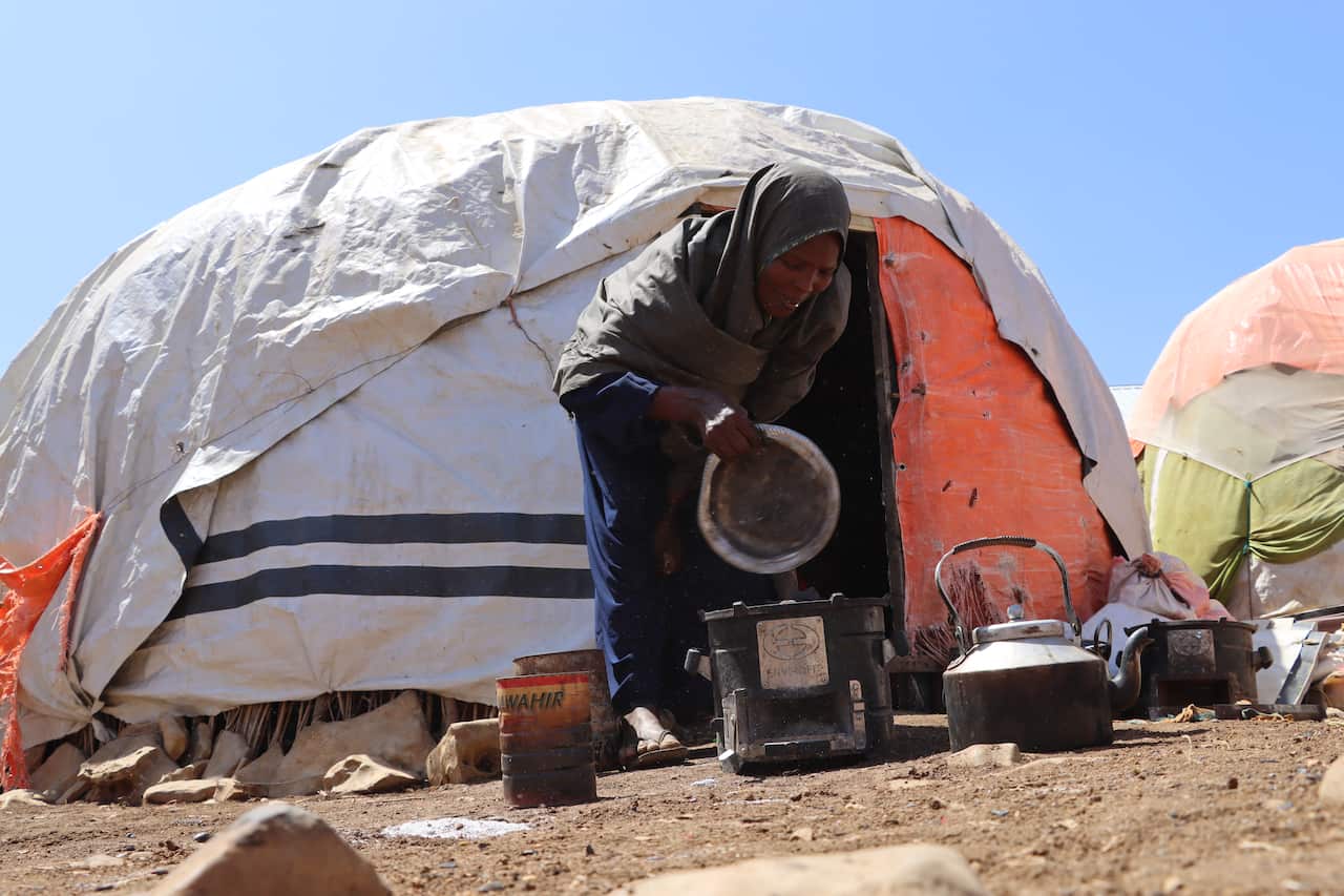 A woman carrying a metal bowl crouches down outside a small white tent 