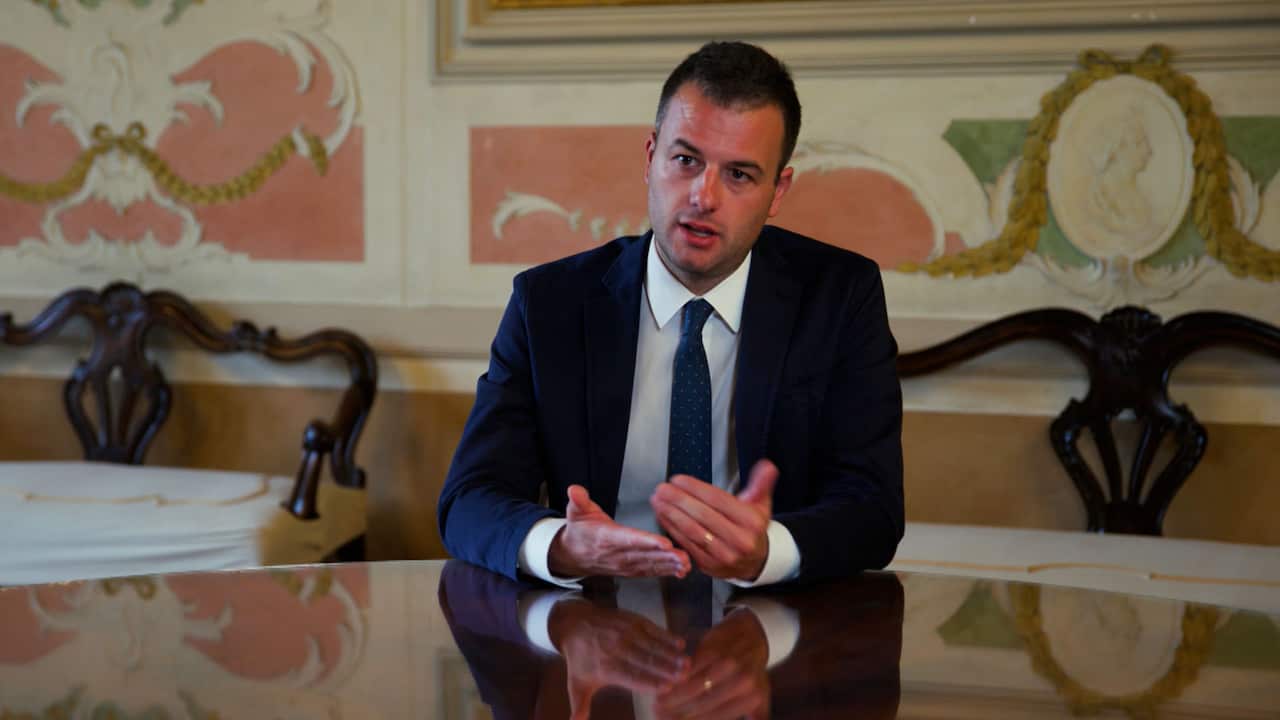 A man in a dark suit, white shirt and a tie is sitting at a table and speaking