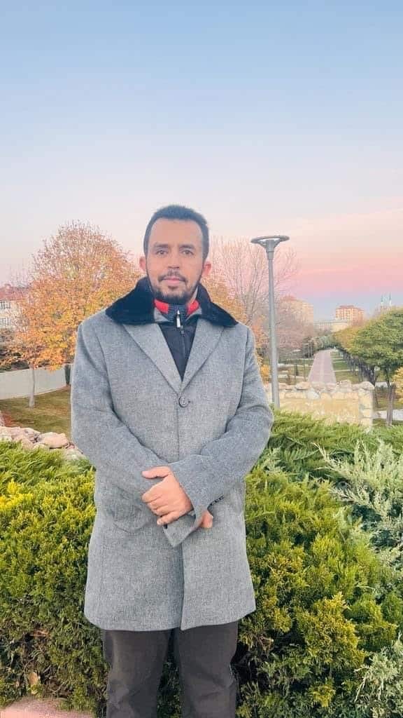 A man stands outdoors in a grey coat, with trees and a landscaped park behind him.