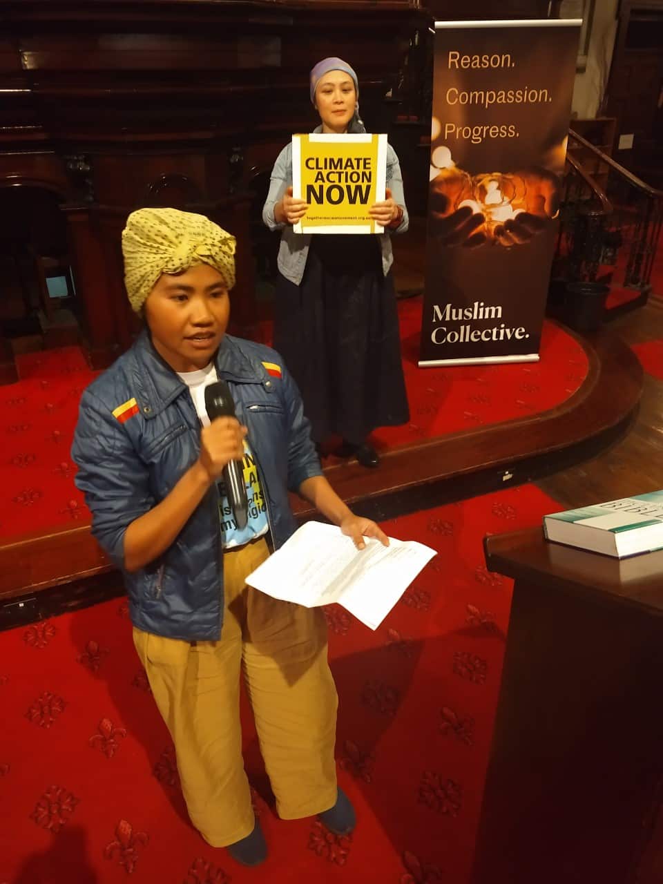 Fahimah holding a microphone and talking indoors. A woman behind her holds a sign which reads: 'Climate action now'