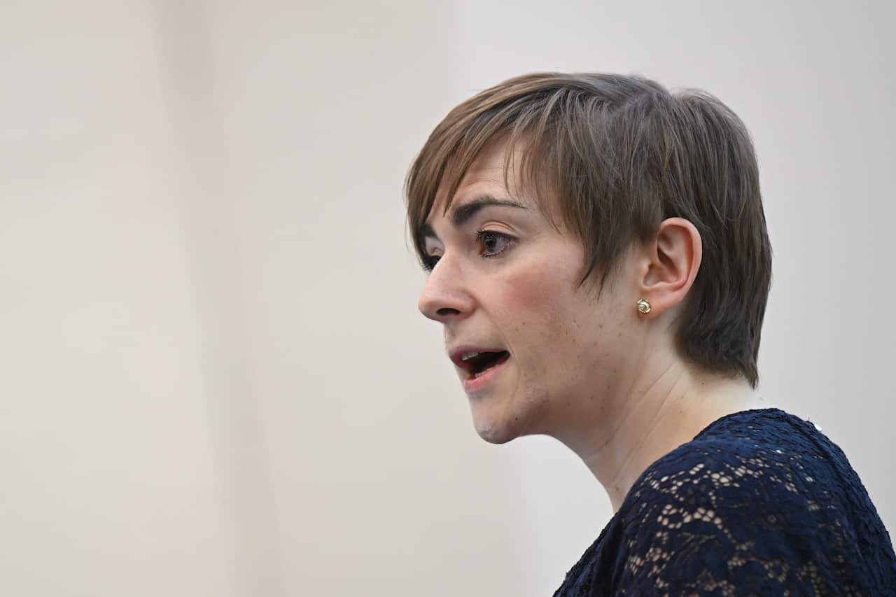 A woman with short brown hair and a dark top, speaking.
