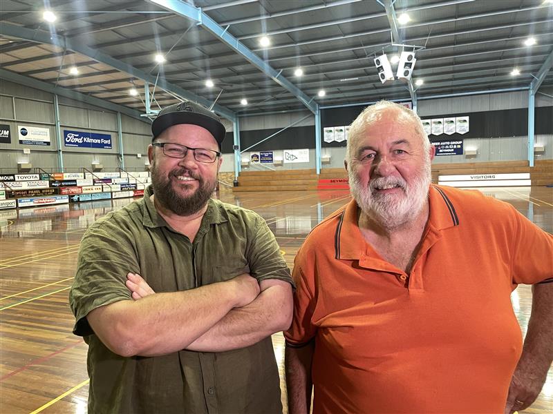 Two white men, one older and one younger, standing beside each other in an indoor sports centre.