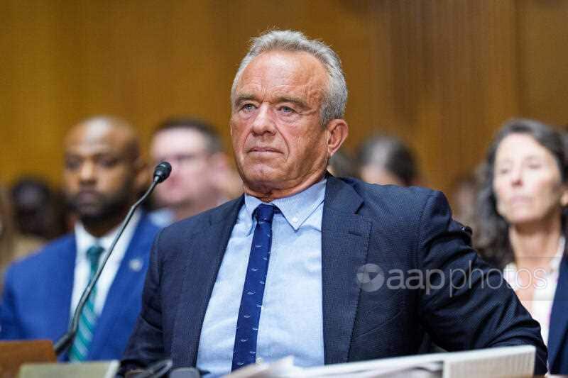 A man in a dark blue suit sits behind a desk with a microphone. There are rows of people seated behind him