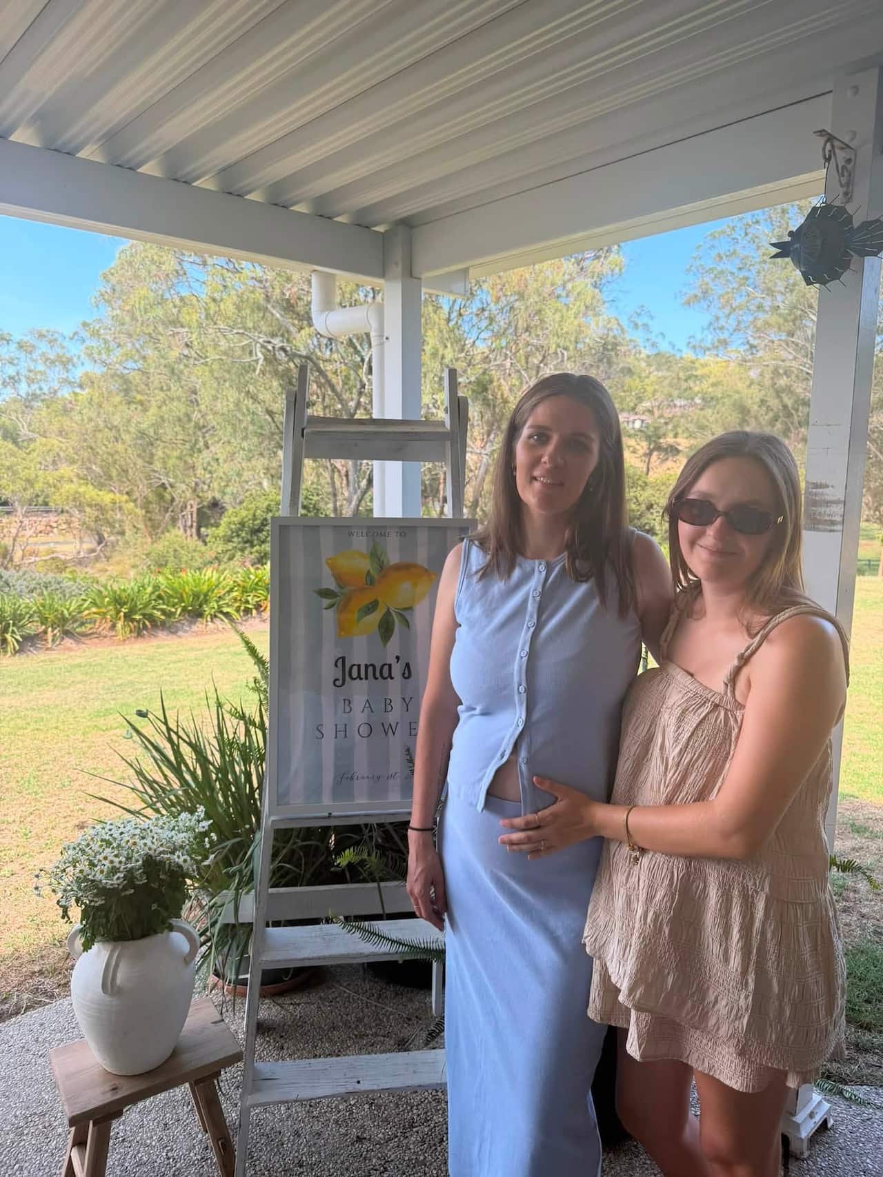 Two women stand together smiling on a covered veranda, next to a welcome sign featuring a lemon illustration that reads "Jana's Baby Shower."