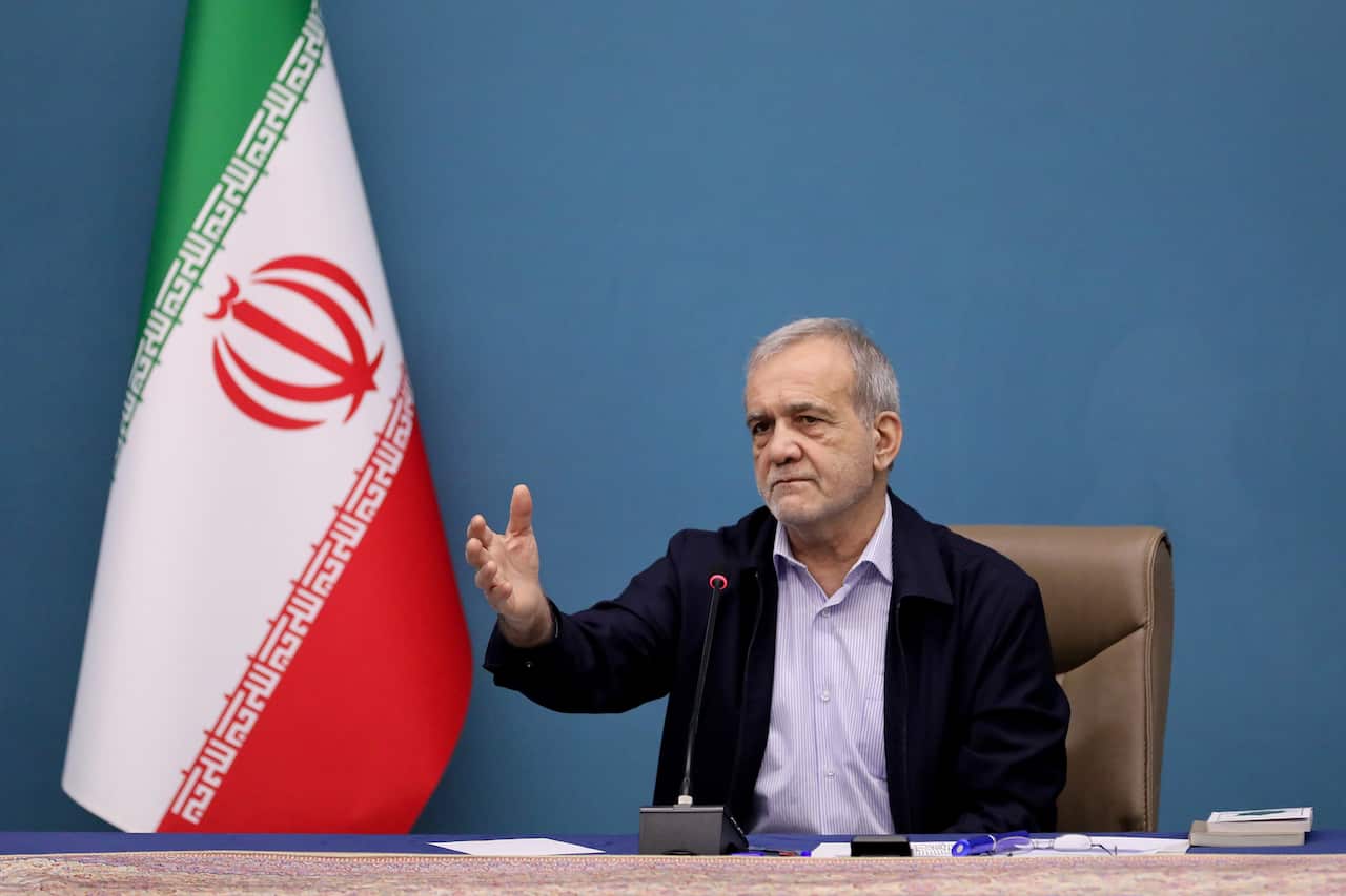 An older Persian man sitting at a desk with his right arm in front of him. An Iranian flag is beside him. 