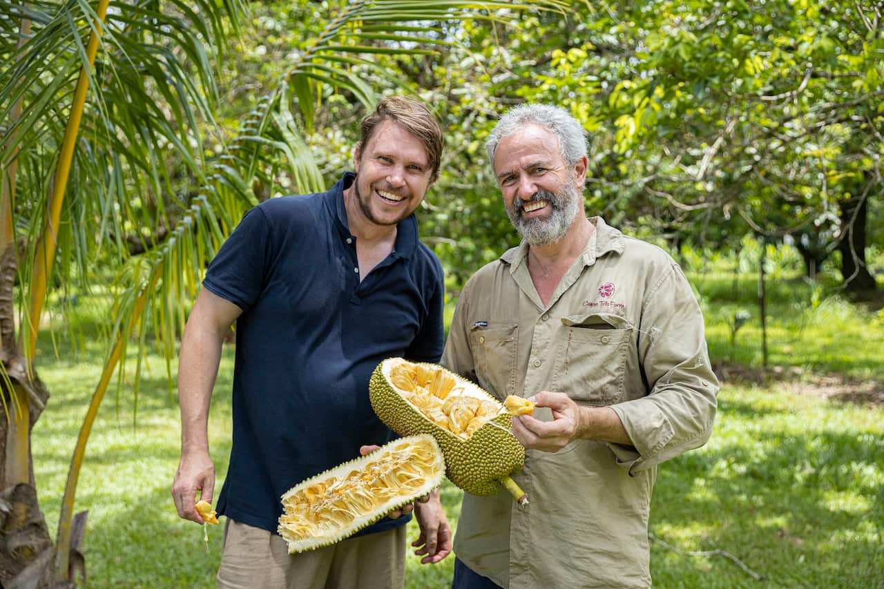 Two men stand in a lush green garden, one of them is holding a very big tripical fruit. 