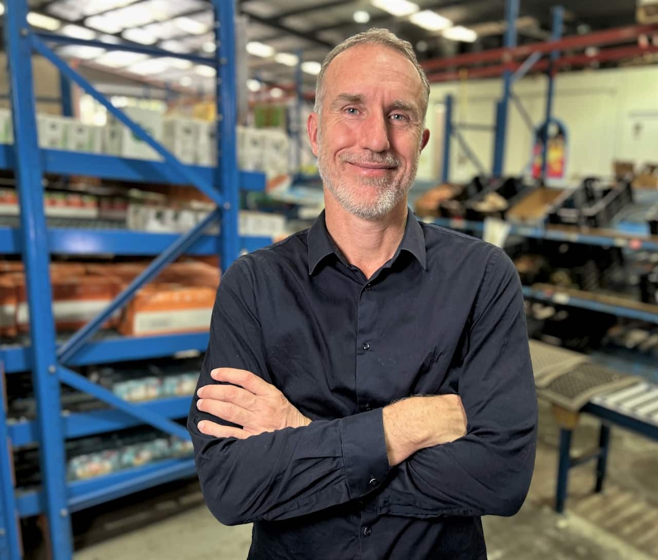 A man standing with arms folded wearing a blue shirt in a warehouse.