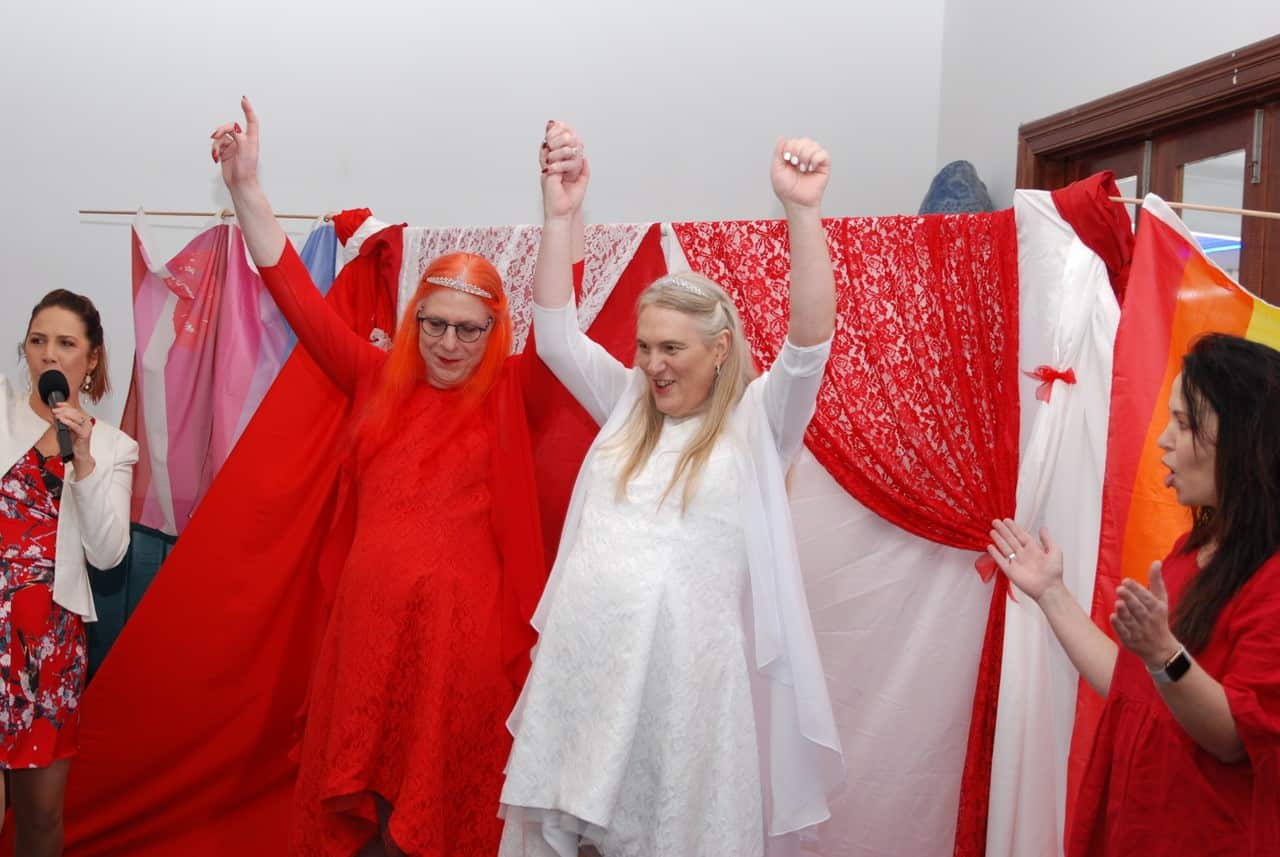 Two women raise their hands inside. One is wearing a red dress, the other a white dress. There is a woman to each side of them cheering.