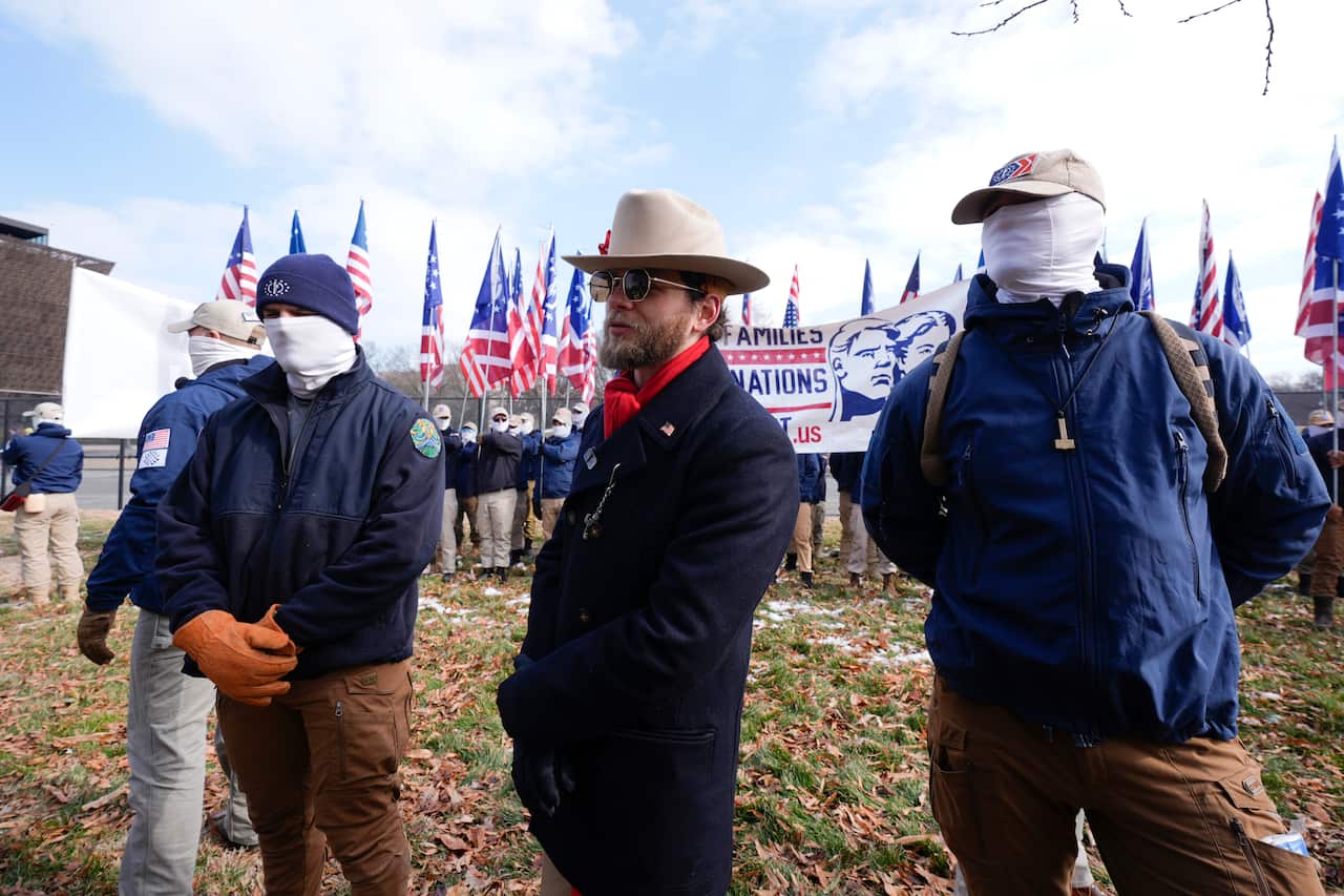 A man in a cowboy hat and dark blue jacket stands between two men with white fabric obscuring their faces.