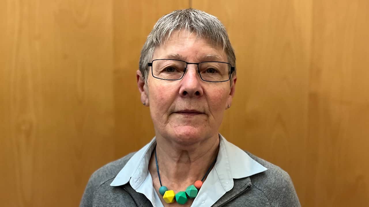 a closeup of a middle aged woman with short grey hair and glasses in front of a timber background