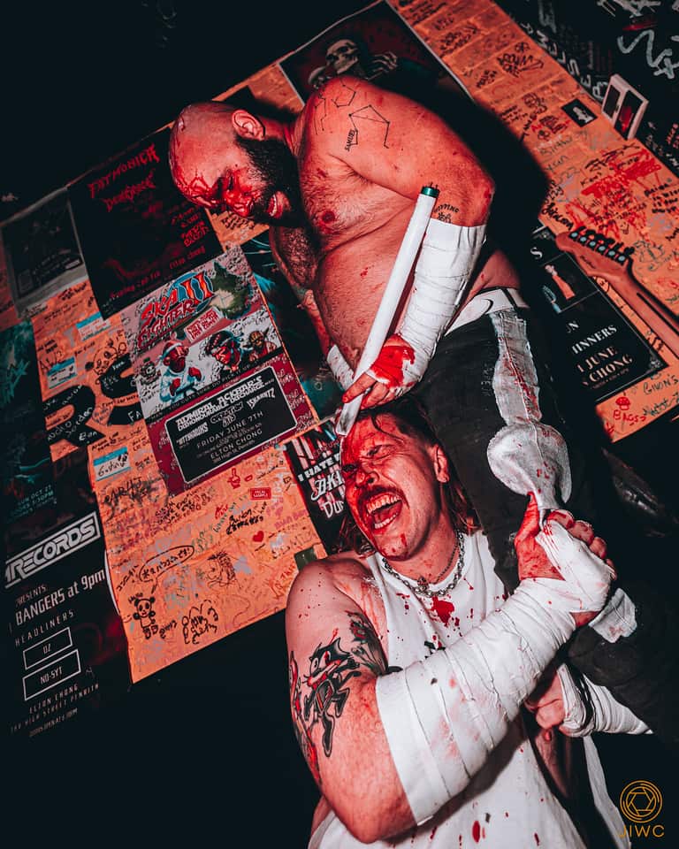 Two men wrestling in front of posters. They're both bleeding in the head. One is smashing a light globe into the other's head. 