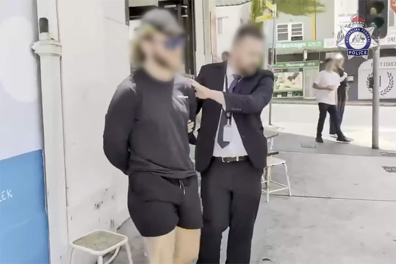 A man in black shorts and t-shirt being arrested by an officer, with both faces blurred.