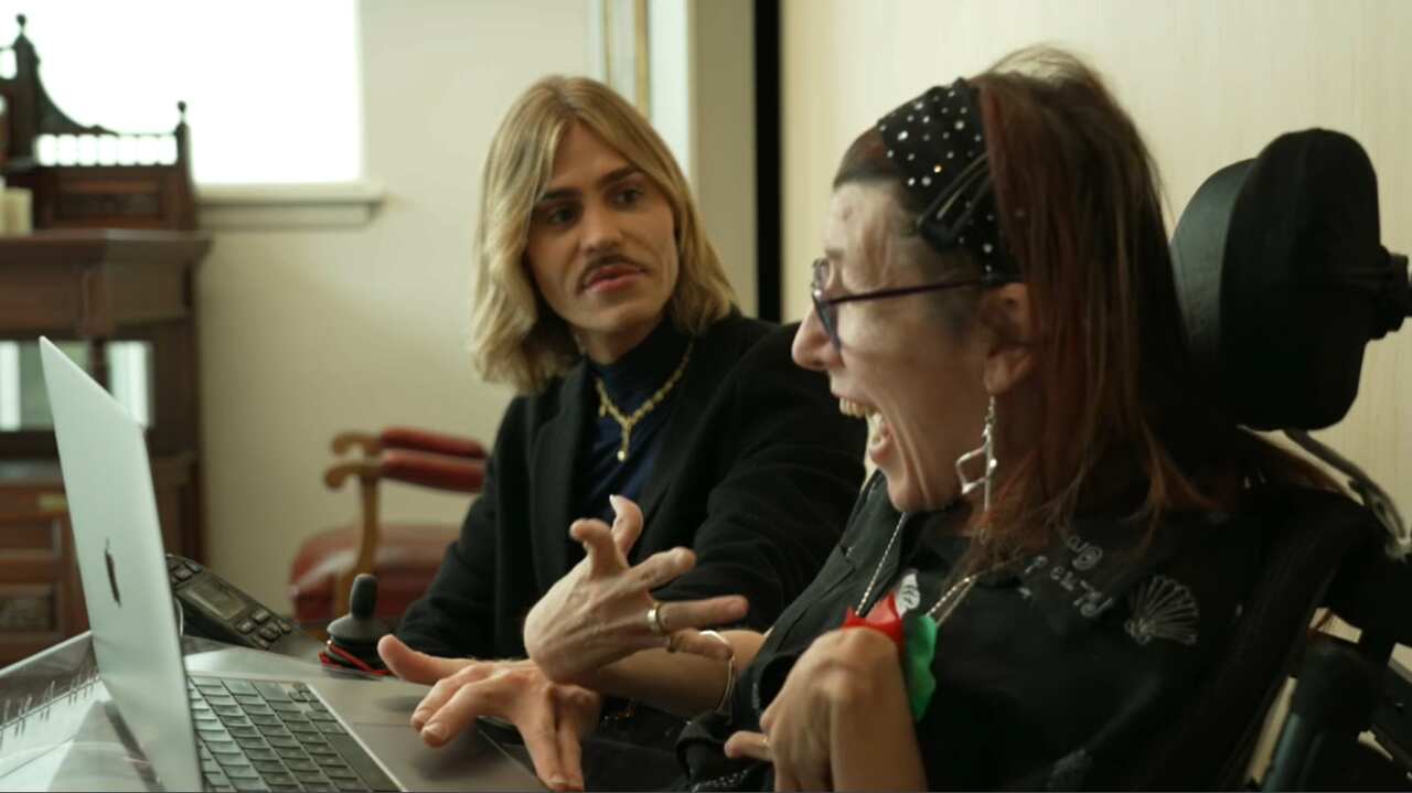 A person with blonde hair and a black top using a laptop alongside a person with red hair sitting in a wheelchair.