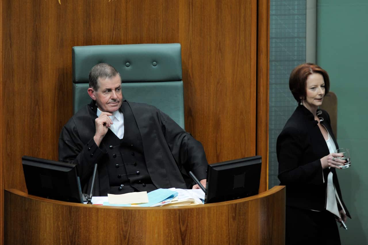 A man dressed in black robes and white shirt sitting at a desk. Julia Gillard, wearing a dark suit and white blouse, is standing to his left