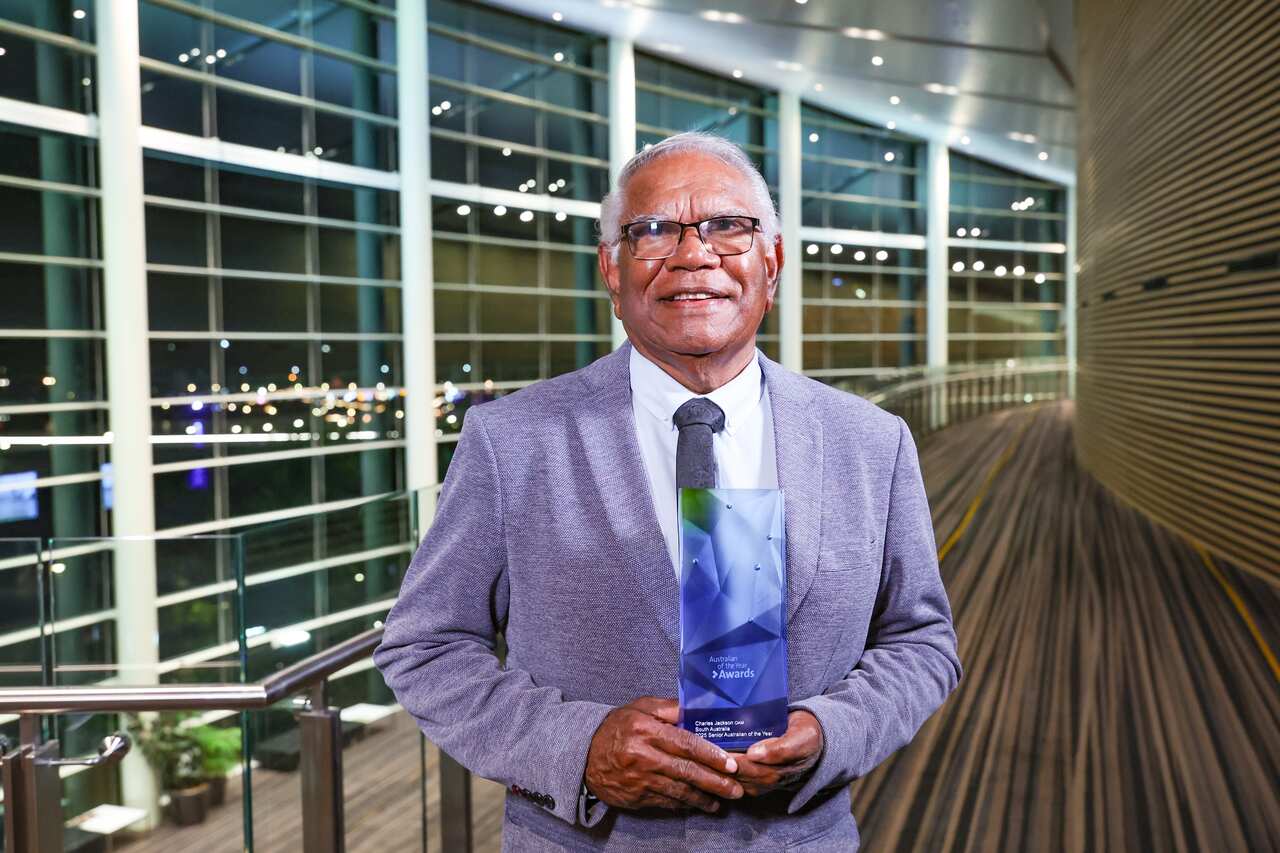 An elderly man wearing a light blue suit and glasses is holding a trophy.