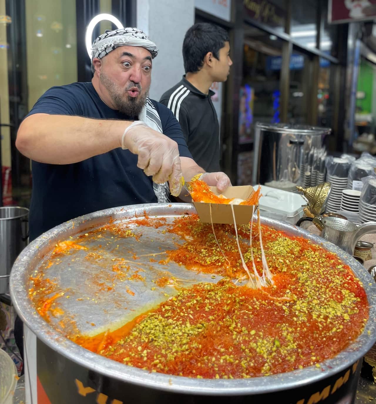 Man wearing kaffeyeh with shocked facial expression pulls knafeh onto container. 