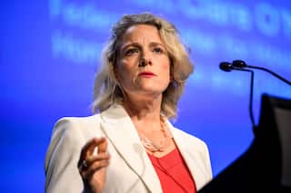 A woman with blonde hair who is wearing a white blazer and orange shirt stands at a lectern with her hand raised.