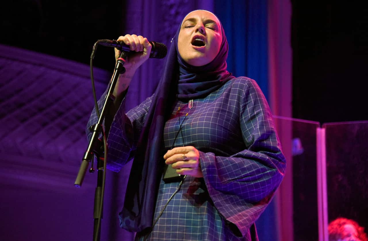 Sinead O'Connor, wearing a long dress and a head scarf, standing and singing into a microphone