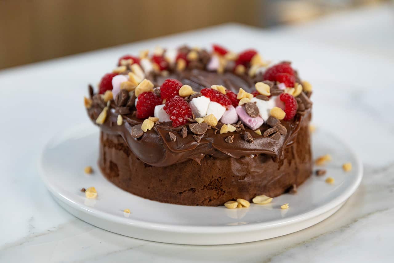 A round chocolate cake sits on a white plate. It is topped with thick swirls of chocolate cing, topped with pices of marshmallow, raspberry, nuts and chocolate. 