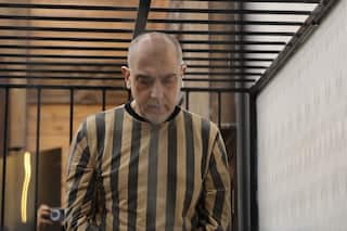 Former Syrian regime official Brigadier General Atef Najib inside a large steel cage, wearing a mustard and black striped prison uniform.