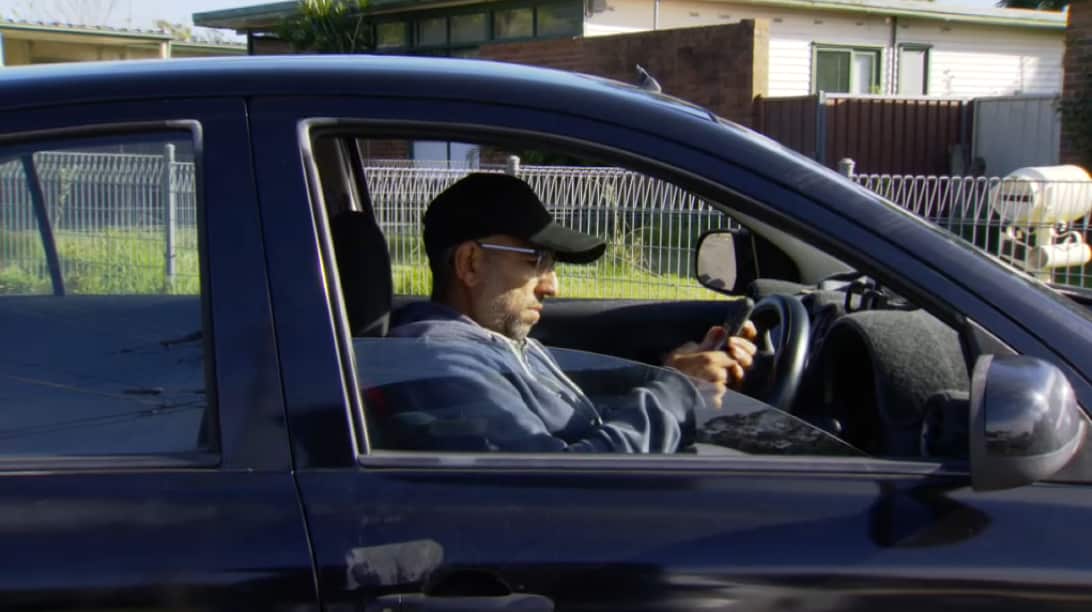 A man wearing a cap is using a phone inside a parked blue sedan.